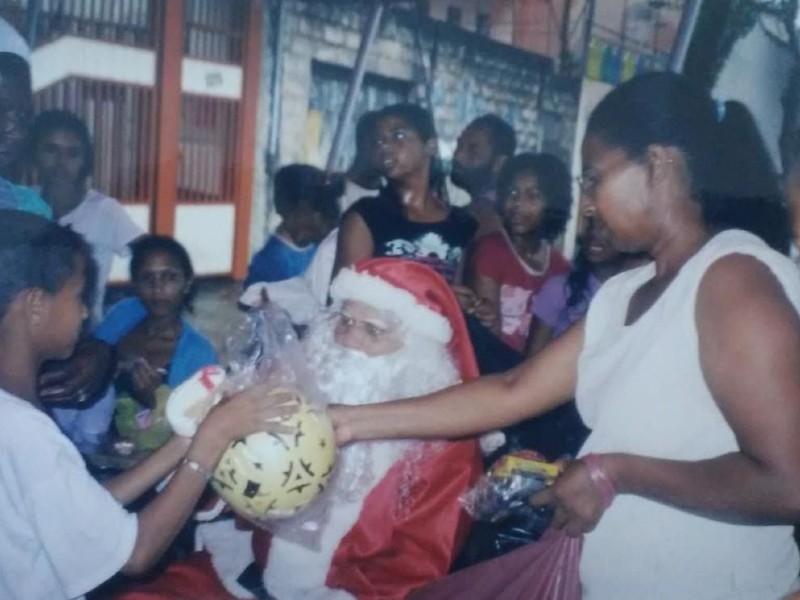 A imagem mostra adultos e crianças em torno de um homem vestido de Papai Noel que está sentado e Rita de Cássia, ao lado em pé, ajudando-o a entregar uma bola de presente para um menino